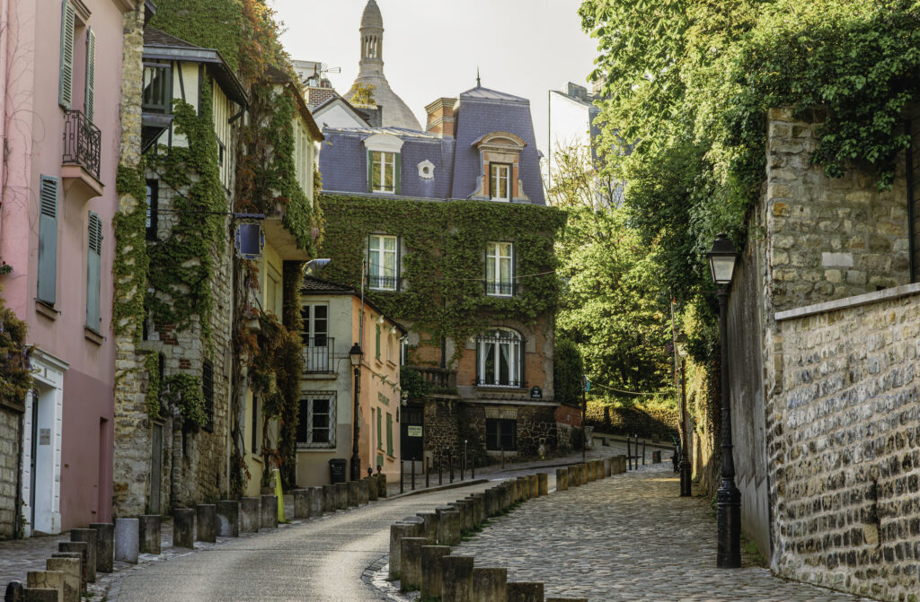 A picturesque scene unfolds in the heart of Montmartre, Paris, as Rue de l'Abreuvoir showcases its timeless charm. The colorful houses line the empty street, radiating with warmth under the sun's embrace on a tranquil spring or summer day. This historic neighborhood epitomizes the romance and allure of the City of Light