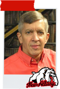 Portrait of a man in a red button-down shirt, in a room with bookshelves in the background; New Albany bulldog logo in the bottom right corner.