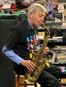 Older man with gray hair blowing into a saxophone in a cluttered music studio filled with books, papers, and equipment.